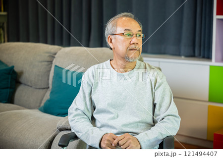 Asian senior man disabled sitting alone on wheelchair in living room looking out window at home and thinking, lonely elder sad old man retirement memory loss or nostalgia thought, feel depressed 121494407