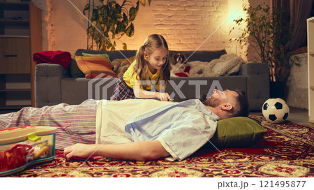 Young girl playing with her father, pretending to be a doctor, using stethoscope. Cute family moment in cozy living room at home in the evening 121495877