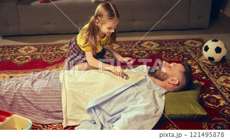 Little girl checks her father health with toy medical tools, playing in cozy living room at home. Pretending to be a doctor 121495878