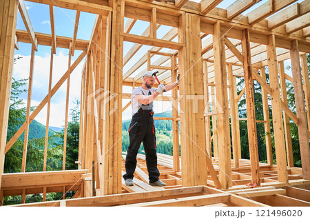 Carpenter constructing wooden frame, two-story house. Bearded man hammering nails with hammer, dressed in protective helmet and work coveralls. Concept of environmentally-friendly modern construction. 121496020