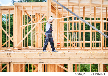 Carpenter constructing wooden-framed house near. Man holds large beam on his shoulder and is dressed in work clothes and helmet. Modern and environmentally-friendly construction concept. Carpenter constructing wooden-framed house near. Man holds large beam on his shoulder and is dressed in work clothes and helmet. Modern and environmentally-friendly construction concept. 121496021