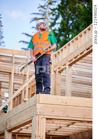 Carpenter building wooden frame two-story house. Bearded man in glasses holding a level, wearing protective helmet, overalls and orange vest. Concept of modern ecological construction. 121496024