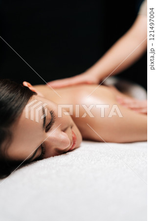 Closeup vertical shot of carefree woman client relaxing during back massage in spa salon with dark interior. Professional massage therapist providing therapeutic back massage to relaxed female. 121497004