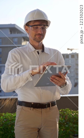 Male construction engineer wearing white shirt and helmet is using digital tablet while inspecting a building site at sunrise, front and vertical view . Architecture concept Male construction engineer wearing white shirt and helmet is using digital tablet while inspecting a building site at sunrise, front and vertical view . Architecture concept 121502023