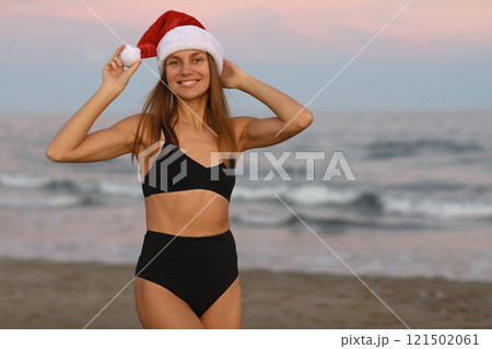 Attractive smiling young woman wearing Santa Claus hat and black bikini on seashore looks at camera. Christmas and New year vacations. Celebrating winter holidays abroad at warm tropical destination. Attractive smiling young woman wearing Santa Claus hat and black bikini on seashore looks at camera. Christmas and New year vacations. Celebrating winter holidays abroad at warm tropical destination. 121502061