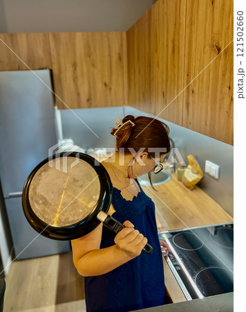 A young girl in the kitchen near an electric stove holds a frying pan in her hands and tries to figure out the equipment A young girl in the kitchen near an electric stove holds a frying pan in her hands and tries to figure out the equipment 121502660