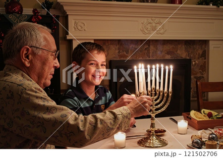 Side view portrait of smiling Jewish boy with grandfather lighting menorah candles during Hanukkah celebration in cozy home setting copy space 121502706