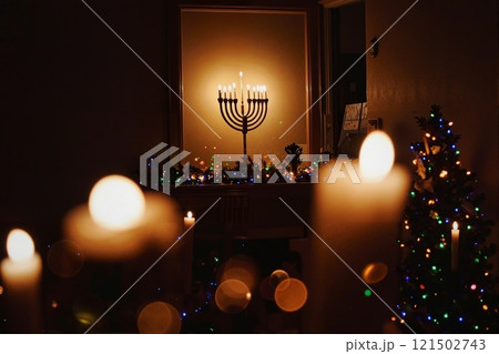 Background image of menorah candles on mantlepiece glowing brightly in dark room with twinkling Christmas lights at night in cozy home setting copy space 121502743