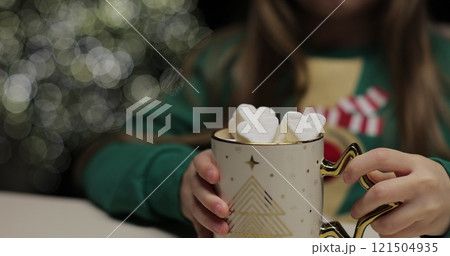 Close-up of little girl in a green Christmas sweater drinks cocoa with marshmallows in the kitchen on glowing Christmas tree background. Christmas mood. 121504935