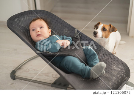 A dog sits next to a cute three-month-old boy dressed in a blue overalls in a baby lounger. 121506075