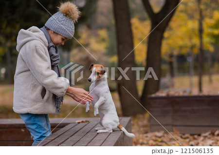 Caucasian girl holding a dog by the paws for a walk in the autumn park. Jack Russell Terrier stands on its hind legs on a bench. 121506135