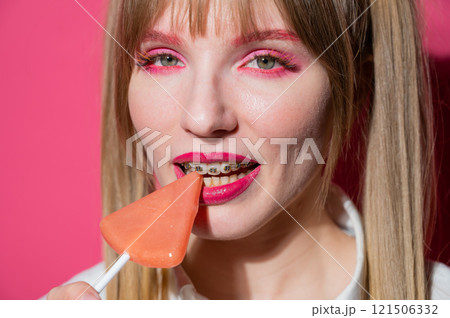 Portrait of a young woman with braces and bright makeup eating a lollipop on a pink background. 121506332