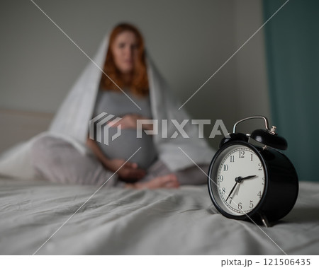 Caucasian pregnant woman sits on the bed and suffers from insomnia. Alarm clock in the foreground. Caucasian pregnant woman sits on the bed and suffers from insomnia. Alarm clock in the foreground. 121506435