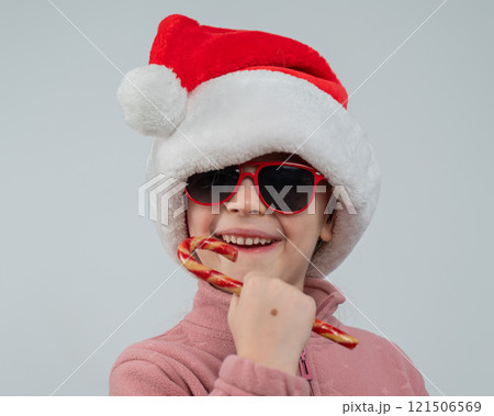 Portrait of a cute Caucasian girl in a Santa Claus hat and sunglasses eating a lollipop on a white background. Portrait of a cute Caucasian girl in a Santa Claus hat and sunglasses eating a lollipop on a white background. 121506569