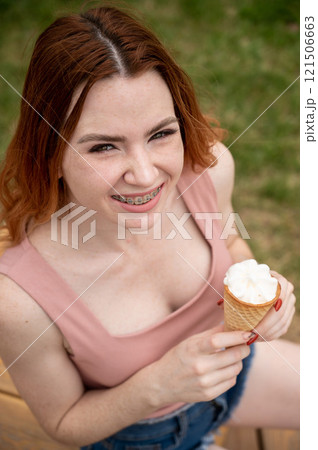 Young beautiful red-haired woman smiling with braces and eating an ice cream cone outdoors. Vertical photo. 121506663