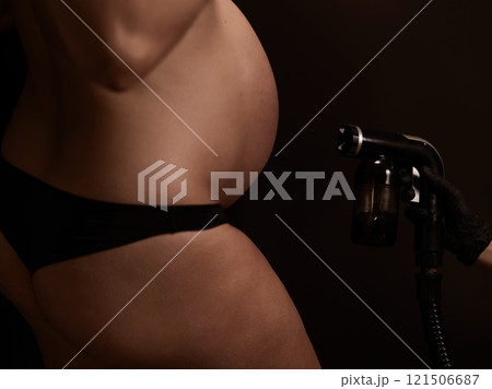 Close-up of the belly of a pregnant woman undergoing an instant tanning procedure. The master applies bronzer with a spray. 121506687
