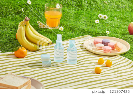 A blue skin care set displayed on a white and yellow striped rug on green lawn, surrounded by some fresh fruits, food and drink for picnic meal. High angle, empty space for design 121507426