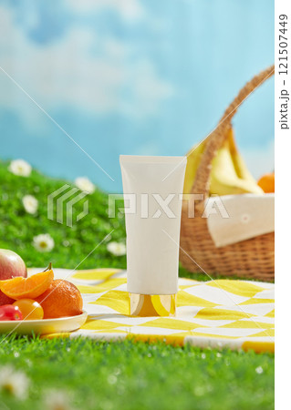 Close-up shot of an unlabeled white tube, placed in the center on a picnic mat, next to a dish filled with some fresh fruits and a basket. A picnic setting with a blue sky backdrop and green grass. Close-up shot of an unlabeled white tube, placed in the center on a picnic mat, next to a dish filled with some fresh fruits and a basket. A picnic setting with a blue sky backdrop and green grass. 121507449