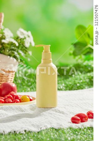 A white picnic mat is laid out in a verdant meadow, along with a daisy basket, a plate with an apple and some tomatoes, and the front of a yellow, unbranded bottle. Cosmetics advertising photo. 121507476