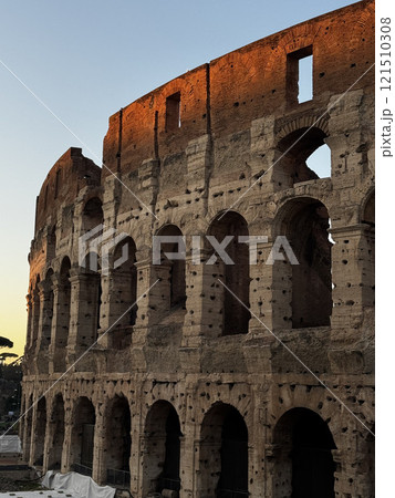 Golden light bathes the ancient Colosseum in Rome during sunset, showcasing its remarkable stone structure and arches. Visitors marvel at this iconic landmark's history and grandeur. 121510308