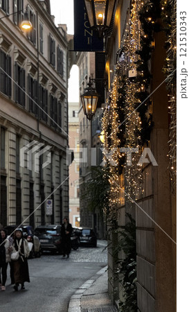 Pedestrians stroll through a narrow Roman alley during a winter evening, while festive lights illuminate the historic buildings and create a warm, inviting atmosphere for the season. 121510343