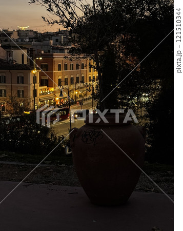 A quaint hillside offers a beautiful view of a bustling city street as twilight falls. Streetlights twinkle and a clay jar rests peacefully in the foreground, capturing the evening's ambiance. 121510344