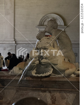 Visitors admire the detailed stone fountain adorned with figures and flowing water in a lively public square illuminated by soft lighting during the evening. 121510354
