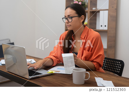 With her laptop, a woman pays household utility bills online, clutching payment receipts for organized financial management at home 121512303