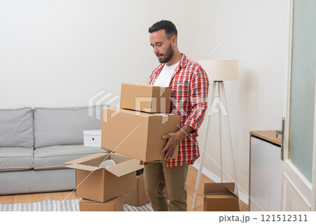 Young man, Caucasian, moves into his new home, standing in the living room with a heap of cardboard relocation boxes, marking the transition Young man, Caucasian, moves into his new home, standing in the living room with a heap of cardboard relocation boxes, marking the transition 121512311