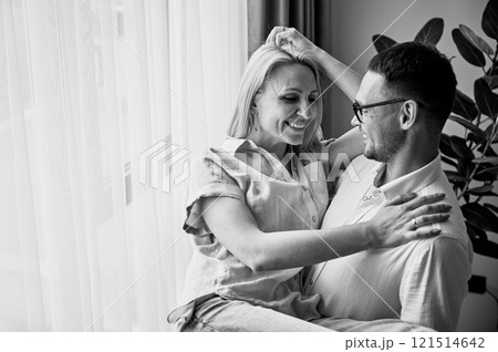 Happy man holding his wife in his arms at home. Lovely couple smiling at each other. Sweet couple looking at each other in casual clothes on light background. Black and white image. Happy man holding his wife in his arms at home. Lovely couple smiling at each other. Sweet couple looking at each other in casual clothes on light background. Black and white image. 121514642