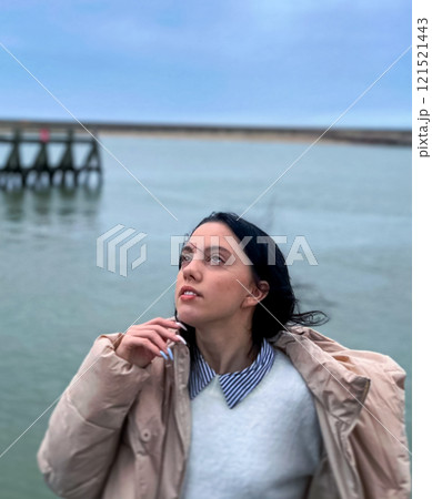 Portrait of a beautiful young woman in a winter jacket standing on a wooden pier in front of the rainy clouds on the background in the Normandy, France. 121521443