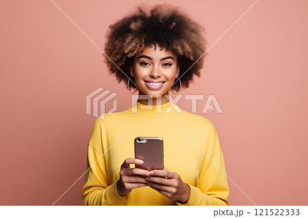 A cheerful woman with curly hair holding a smartphone, dressed in a vibrant yellow sweater against a pink background. 121522333
