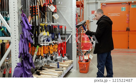 Customer selecting sledgehammer, testing weight, comparing sizes near shopping basket amid hardware store displays of construction tools and professional equipment. Translation: Sale Customer selecting sledgehammer, testing weight, comparing sizes near shopping basket amid hardware store displays of construction tools and professional equipment. Translation: Sale 121522487