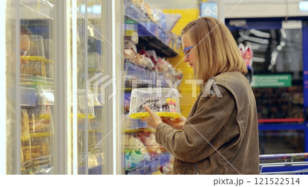 Woman perusing diverse dessert options in a supermarket refrigerated display, selecting a packaged cake and highlighting consumer choices in grocery shopping 121522514