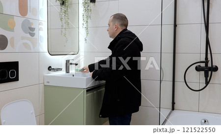 Man washing hands in modern bathroom, green vanity unit, illuminated mirror, black fixtures highlighting contemporary design aesthetic Man washing hands in modern bathroom, green vanity unit, illuminated mirror, black fixtures highlighting contemporary design aesthetic 121522550