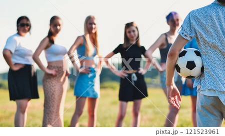 Young man is holding a soccer ball behind his back while standing in a field with a group of women. 121523961