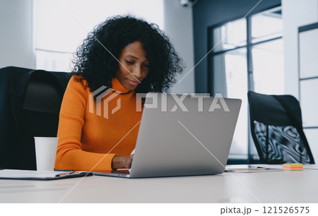 Concentrated professional woman in a bright orange top engages with laptop in a modern office, embodying focused productivity. 121526575