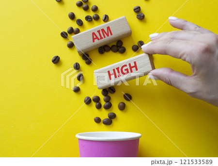 Aim High symbol. Concept word Aim High on wooden blocks. Beautiful yellow background with coffee cup. Businessman hand. Business and Aim High concept. Copy space Aim High symbol. Concept word Aim High on wooden blocks. Beautiful yellow background with coffee cup. Businessman hand. Business and Aim High concept. Copy space 121533589