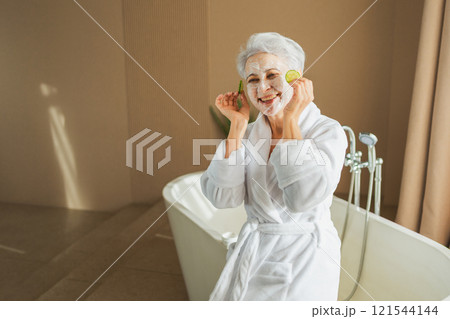 Mature adult old woman with nourishing anti age face cream mask holding cucumber slices. Senior woman with white mask creme on skin gets facial treatment daily beauty routine. Skin care aging beauty Mature adult old woman with nourishing anti age face cream mask holding cucumber slices. Senior woman with white mask creme on skin gets facial treatment daily beauty routine. Skin care aging beauty 121544144