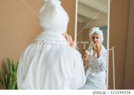Mature adult old woman looking at mirror applying nourishing anti age face cream doing daily beauty routine. Senior woman with white mask creme on skin gets facial treatment Skin care aging skincare Mature adult old woman looking at mirror applying nourishing anti age face cream doing daily beauty routine. Senior woman with white mask creme on skin gets facial treatment Skin care aging skincare 121544156