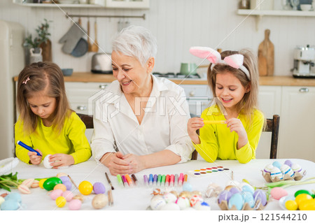 Happy Easter. Two little girls sisters twins grandmother painting eggs enjoying time together. Happy family grandma granddaughters child kids preparing for Easter. Spring Christian festival tradition Happy Easter. Two little girls sisters twins grandmother painting eggs enjoying time together. Happy family grandma granddaughters child kids preparing for Easter. Spring Christian festival tradition 121544210