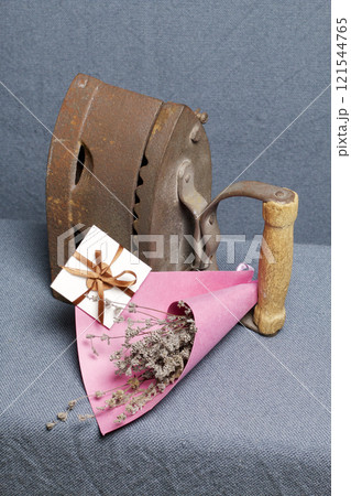 Old iron, heated by hot coals. Located on gray fabric. Nearby are wicker baskets, a bouquet of dried flowers and a greeting card. Old iron, heated by hot coals. Located on gray fabric. Nearby are wicker baskets, a bouquet of dried flowers and a greeting card. 121544765