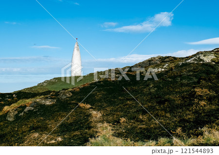 A modern, white tower-like object is situated on a hill. The hill's surface is uneven, with exposed rocks and patches of grass. The day is sunny. 121544893