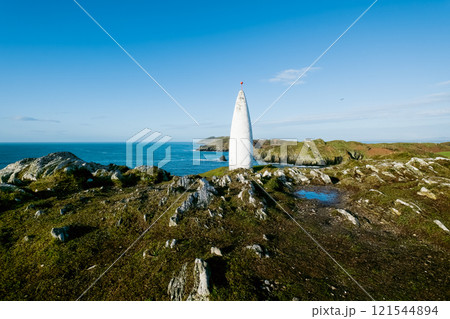 Ocean view. A tall, slender, white cone-shaped tower stands prominently on a rocky hill overlooking a tranquil ocean. Ocean view. A tall, slender, white cone-shaped tower stands prominently on a rocky hill overlooking a tranquil ocean. 121544894