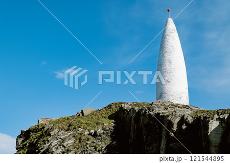 This photo depicts a tall, white, cylindrical structure situated on a cliff top overlooking a clear blue expanse of sky. 121544895