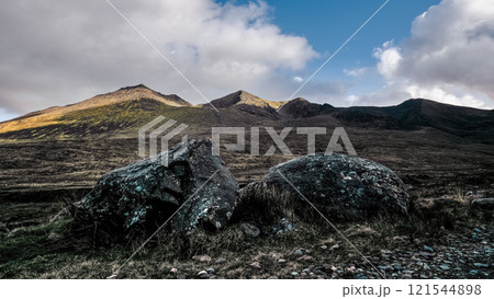 Foreground displays two dark, rounded rocks amidst sparse vegetation. Background shows a mountainous terrain under a blue sky with clouds. 121544898