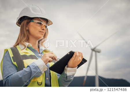 An experienced female engineer is inspecting a wind turbine site, using her tablet to assess the equipments functionality and ensure compliance with strict sustainability standards An experienced female engineer is inspecting a wind turbine site, using her tablet to assess the equipments functionality and ensure compliance with strict sustainability standards 121547486