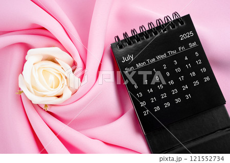 July 2025 desk calendar black color and rose on pink textile. 121552734