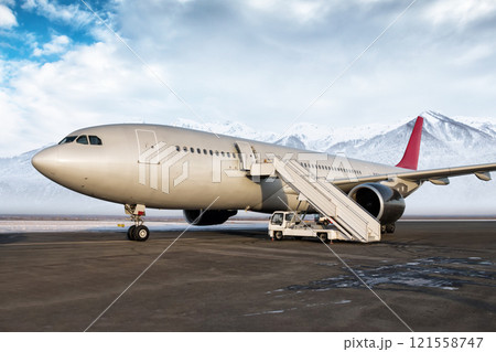 Wide body passenger airplane with boarding steps at the airport apron on the background of high picturesque snow capped mountains Wide body passenger airplane with boarding steps at the airport apron on the background of high picturesque snow capped mountains 121558747