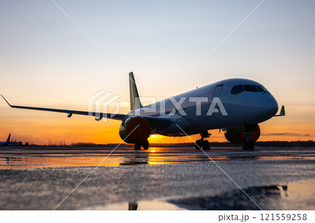 Modern passenger jetliner on the airport apron against the backdrop of a picturesque sunset with reflection in a puddle Modern passenger jetliner on the airport apron against the backdrop of a picturesque sunset with reflection in a puddle 121559258